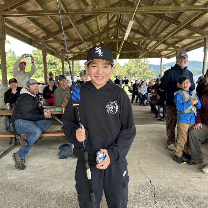 A smiling boy wearing a black track suit holds a fishing rod with a group seated behind him in a picnic structure. 